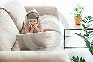 With headphones in place, a sweet little girl relaxing on a beige sofa, engrossed in her laptop screen. Concept: technology- photo
