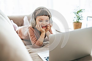 With headphones on, a sweet little girl, relaxing on a sofa, engrossed in the captivating content playing on her laptop photo