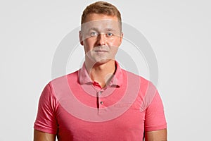 Headshot of attractive handsome man has appealing look, dressed in casual pink t shirt, ooks seriously at camera, over wh photo