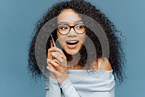 Headshot of attractive young African American woman holds modern cell phone near ear, has nice conversation, focused aside,