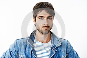 Headshot of attractive young hispanic man with blue eyes and beard in denim jacket over striped t-shirt looking at