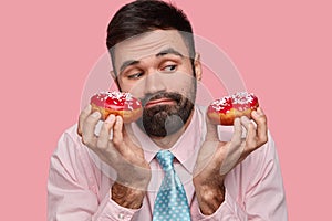 Headshot of attractive young man has puzzled facial expression, holds tasty doughnuts, looks confusingly, has