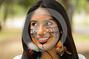 Headshot beautiful Amazonian woman, indigenous facial paint and earrings with colorful feathers, posing happily for
