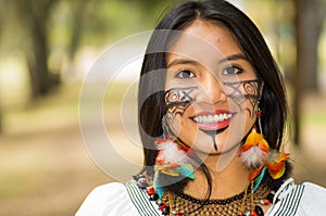 Headshot beautiful Amazonian woman, indigenous facial paint and earrings with colorful feathers, posing happily for photo