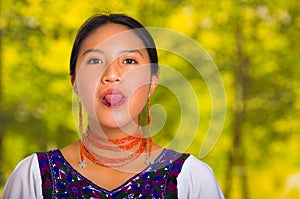 Headshot beautiful young woman wearing traditional andean blouse with red necklace, posing for camera holding tongue out