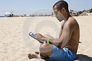 Headshot of black male, feeling happy to spend his free time very well sitting against blue sea and working with tablet.