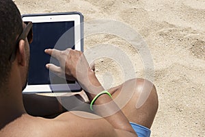 Headshot of black man, sitting on the beach and working with tablet.