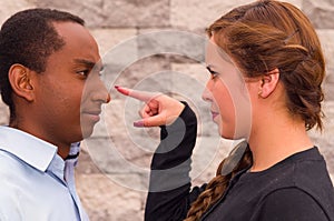 Headshot charming young interracial couple facing each other, she point her finger at his nose, brick wall background