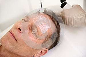 Headshot. Close-up of a handsome mature 50 years old man with closed eyes enjoying beauty treatment in spa salon, while beautician