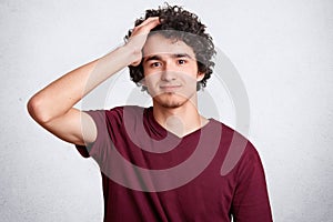 Headshot of handsome youngster, has curly hair, keeps hand on head, wears casual clothes, poses against white background. Joyful t