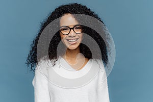 Headshot of lovely curly woman being in good mood, smiles broadly, expresses nice feelings, wears transparent spectacles and white