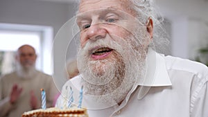 Headshot portrait of bearded happy senior man blowing out candles on cake smiling. Caucasian male retiree celebrating