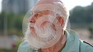 Headshot portrait of bearded old man with brown eyes looking around standing on city street. Close-up of male Caucasian