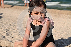 Headshot portrait of cute upset baby girl with wet hair and swimsuit, sitting on the sandy beach on the sea background. Kids