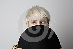 Headshot Portrait of happy blonde girl hides her face behind black hat looking asside at camera. White background