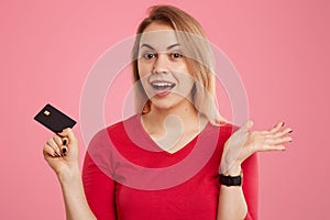 Headshot of positive hesitant light haired European woman dressed in red jumper, holds credit card, has clueless expression, poses
