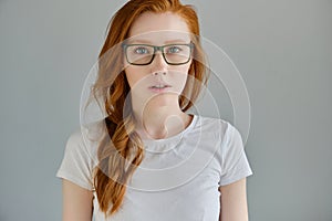 Headshot. Red-haired girl in a white T-shirt and glasses stands on a gray background and looks into the frame.