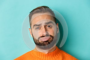 Headshot of sad and gloomy man complaining, pouting and frowning disappointed, standing against turquoise background