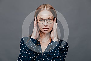 Headshot of serious female teacher keeps hands on head, has self confident facial expression, wears spectacles, polka dot white