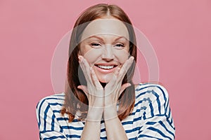 Headshot of smiling young European woman has make up, smiles broadly, keeps hands on cheeks, shows her beauty, expresses good