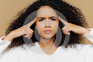 Headshot of stressful Afro American woman keeps index fingers on temples, suffers from headache frowns face, has dissatisfied