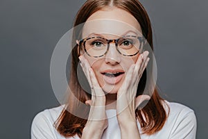 Headshot of surprised young Caucasian lady keeps both palms on her cheeks, looks with amazement, recieves unexpected news, wears