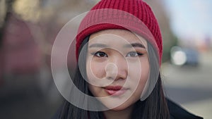 Headshot of young slim charming Asian woman in red hat looking at camera smiling standing outdoors on sunny spring