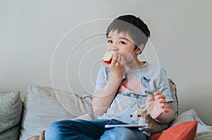 Healhty kid eating an apple while watching TV, Portrait child eating fresh fruit. Happy boy sitting on sofa relaxing at home on