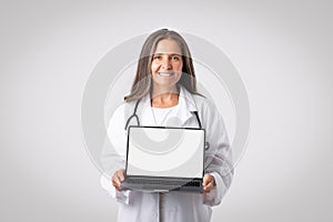 Health app promotion concept. Happy senior woman doctor holding modern pc laptop with black empty screen, mockup