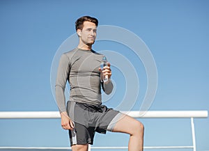 Health and body care. Muscular handsome guy in sportswear, with wireless headphones holding bottle of water on blue sky