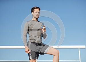 Health and body care. Muscular handsome guy in sportswear, with wireless headphones holding bottle of water on blue sky photo