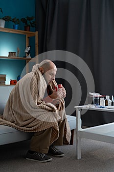 health, cold and people concept. Sad sick man drinking hot tea at home sitting on sofa wrapping in plaid