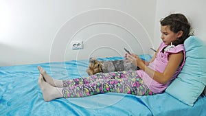Health, technology and beauty concept - little girl with smartphone playing in bed and dog