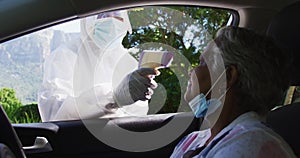 Health worker in protective clothes measuring temperature of african american senior woman in car