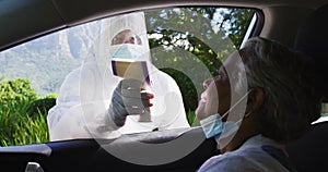 Health worker in protective clothes measuring temperature of african american senior woman in car