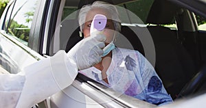 Health worker in protective clothes measuring temperature of african american senior woman in car