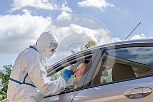 A healthcare worker completely covered in a white protective suit and glasses operates a check swab for Covid-19 to a man in a car
