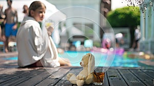 Healthful vitamin juice and rabbit toy on poolside deck with blurred girl drinking sitting at background in towel