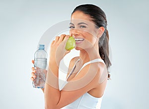 Healthiness is happiness. Studio shot of a fit young woman holding a bottle of water and an apple against a gray