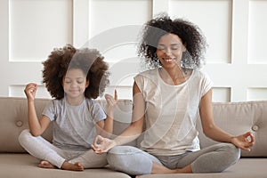 Healthy african mother teaching meditation sit with daughter on sofa