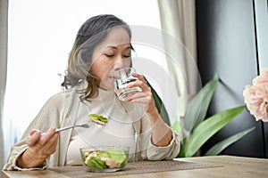 Healthy Asian-aged eating salad and drinking water in her dining room