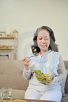 Healthy Asian aged woman enjoys eating salad vegetables mix on sofa in her living room