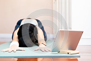 Healthy Asian woman doing yoga child pose stretching her back and upper body with tablet computer in bedroom at home