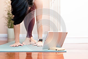 Healthy Asian woman standing cross legged forward fold to stretching hamstrings, and hip flexor with online course from tablet