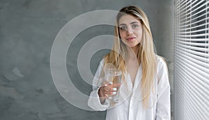 healthy beautiful young woman stands at home near the window holding a glass of water in her hands.