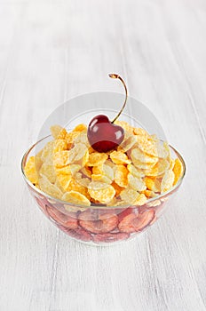 Healthy breakfast in bowl with golden corn flakes, ripe slice cherry on white wood board.