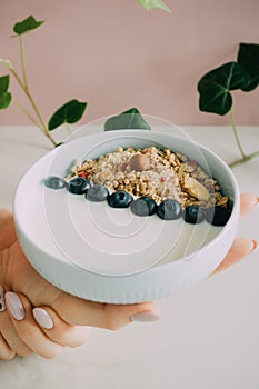 Healthy breakfast bowl with muesli and yogurt in woman hands on pink and marble background.