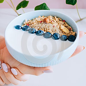 Healthy breakfast bowl with muesli and yogurt in woman hands on pink and marble background.