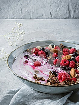 Healthy breakfast bowl with yogurt, granola, muesli, seeds, frozen raspberries, blueberries, nuts in blue ceramics on light