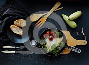 Healthy breakfast: broccoli, cherry tomatoes and fried eggs in a pan, which stands on a wooden stand on a dark background.